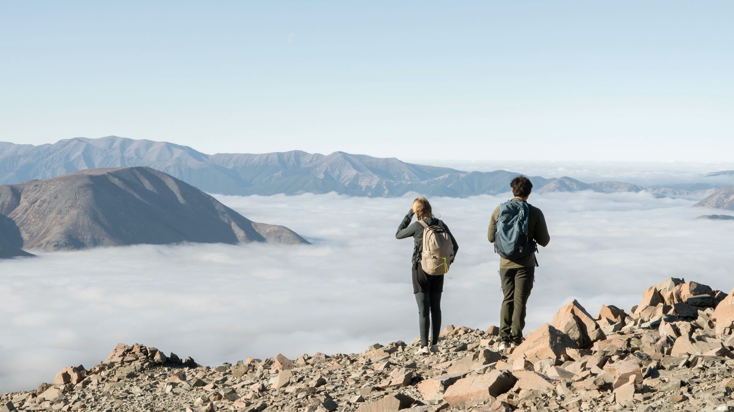 Two people standing on a mountain top with a view of clouds below